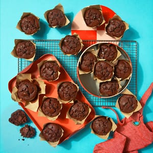 A full case of Chocolate Muffins by Cake Club on a tray and cooling rack with a light blue background.