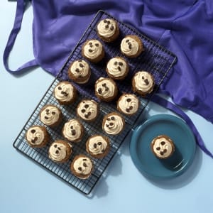 A whole tray of Little Latte mini cakes on a cooling rack, with an individual mini cake on a round blue plate next to a purple apron on a light blue background.