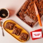 Three Choc Chunk Brownie slices cut and served on a yellow plate beside the remaining brownie tray and a coffee.