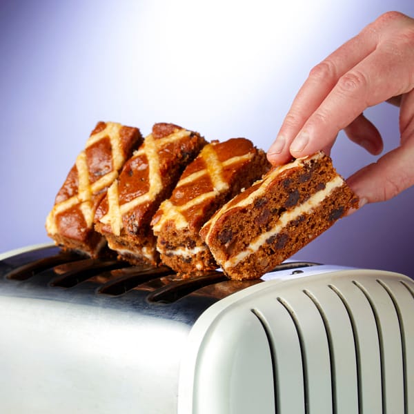 Four slices of Hot Cross Bun Traycake on a white toaster with a hand grabbing a piece with a purple background.