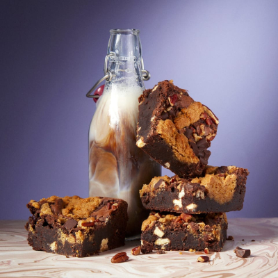Four Cake Club Brookie portions stacked next to a milk bottle filled with creamy chocolate milk in front of a purple background.