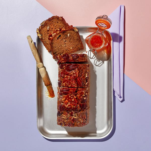 overhead image of banana pecan and rye loaf cake sliced on the silver tray with a pot of apricot glaze on a purple and pink background