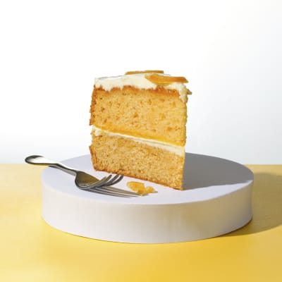 A Lemon and Yuzu slice sitting on a white cake board next to a fork in front of a yellow and white background
